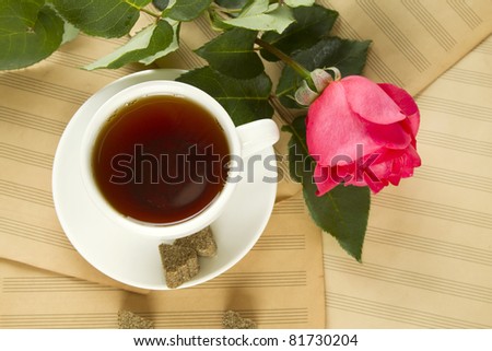 White cup of tea next to sugar and is a beautiful red rose lying on sheets of old paper sheet music