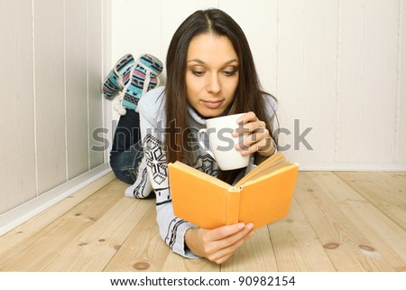 Attractive young woman lying on the floor at home reading a book and drinking coffee