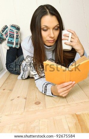 Attractive young woman lying on the floor at home reading a book and drinking coffee