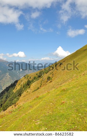 close up of slope of hilly green grass land with backdrop of snow clad peak and blue sky