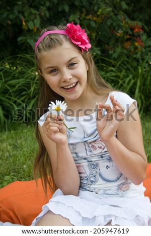 Girl guesses, tearing off petals on camomile, against green summer garden.