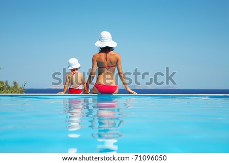 Young mother with baby daughter sitting on the edge of the pool and stare at the sea Young mother with baby daughter sitting on the edge of the pool and stare at the sea