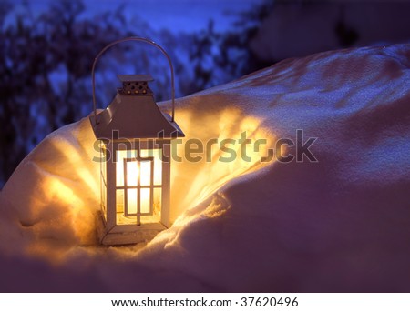 White Christmas lantern in snow, with yellow candle light, on mysterious dark blue background