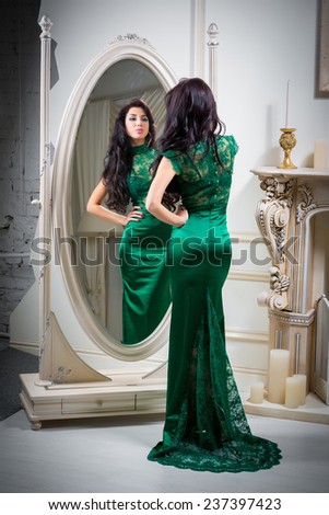 Beautiful sexy young woman in green evening dress is looking at the mirror