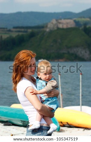 Mother holds in arms her baby on lake background
