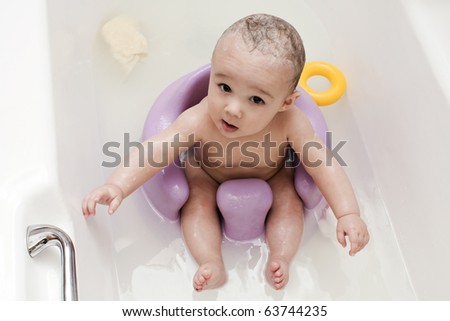 Baby boy bathing in the bath tub and sitting on a purple support chair whilst staring upwards to the camera.