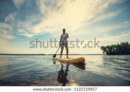 Joyful man is training  SUP board in large river on a sunny morning against a blue sky background . Stand up paddle boarding - awesome active outdoor recreation. Wide angle, backlight.