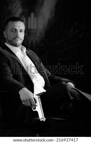 Sophisticated young man agent police killer hitman assassin sitting in a chair holding a gun over dark background black and white portrait