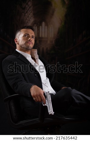 Stylish hispanic young handsome man model mobster spy hitman killer looking up sitting in a chair holding a gun over dark background