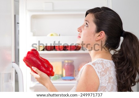 young beautiful brunette woman searching for food in the fridge holding red capsicum