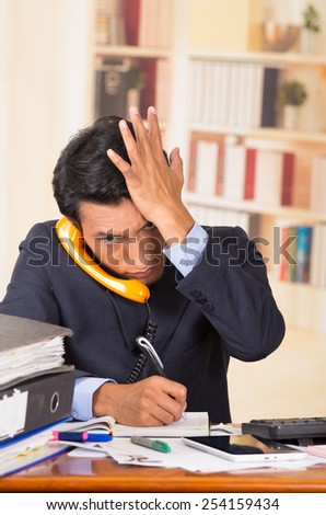 young stressed overwhelmed business man with piles of folders on his desk talking on the phone
