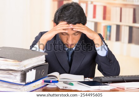 young stressed overwhelmed business man holding head with his hands looking at piles of folders on his desk