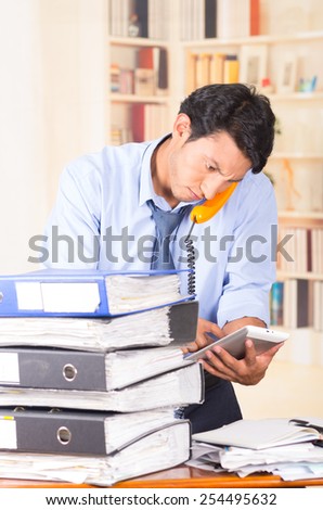 young stressed overwhelmed business man with piles of folders on his desk talking on the phone