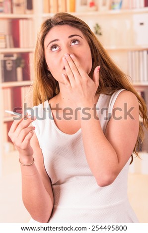 nervous beautiful young girl holding pregnancy test with hand on her mouth gesturing surprise