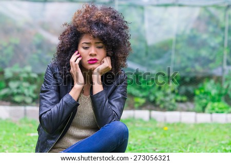 exotic beautiful young girl with dark curly hair using her cell phone sitting in the garden, looking worried