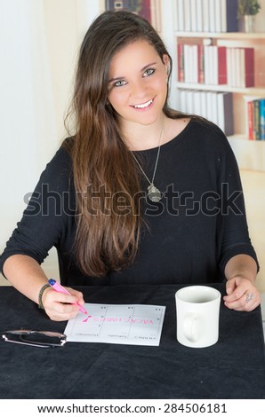 brunette girl sitting by desk with coffee mug and writing on a paper while smiling towards camera, spanish words say friday, saturday, sunday, vacation