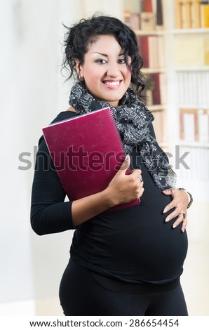 pregnant woman wearing casual clothes at work standing with folder and touching belly