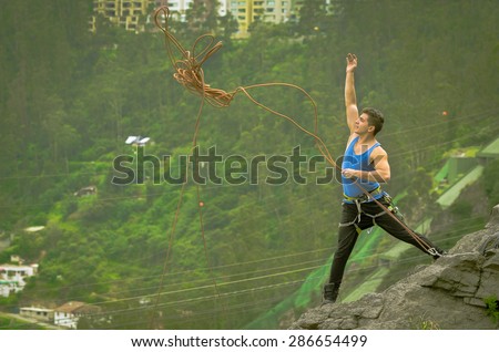 athletic man standing on cliff throwing rope with right arm down mountain