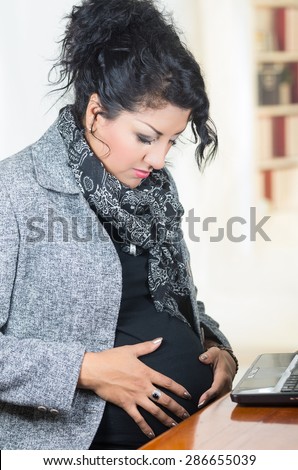 closeup of pregnant hispanic woman wearing casual clothes at work sitting by desk touching belly