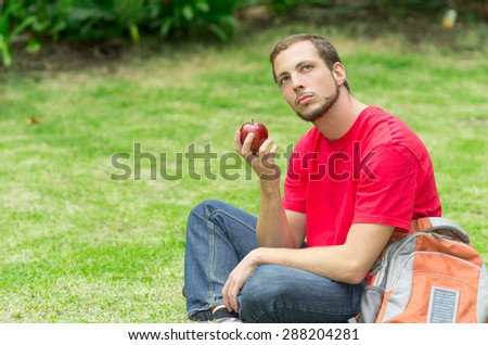 Man wearing jeans and red t-shirt sitting in park holding an apple