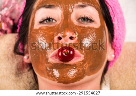 Closeup womans face covered in brown facial treatment cream and looking to camera with cherry on top of mouth