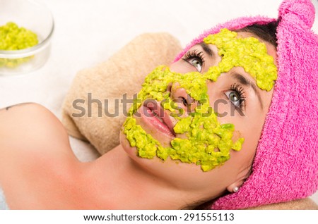 Womans face covered with green cream and eyes open wearing pink towel on head lying down