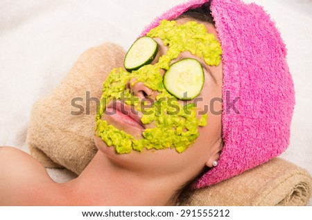 Womans face covered with green cream and cucumber over eyes wearing pink towel on head lying down