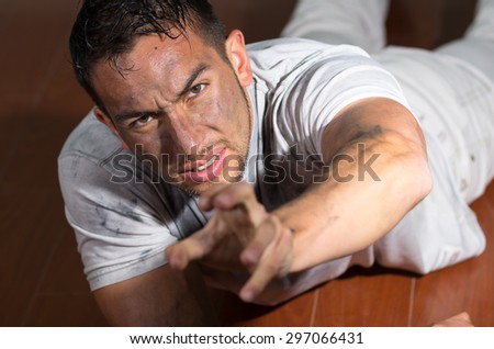 Hispanic man with dirty face and shirt lying on floor looking desperately to camera reaching left hand towards lens.