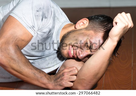 Hispanic man with dirty face and shirt on floor locking burying head inside left arm as in pain.