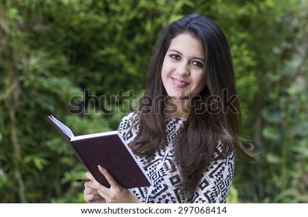 Hispanic brunette in park environment wearing formal clothing holding book open while smiling mouth closed towards camera.