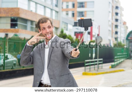 Man wearing formal clothing posing with selfie stick in urban environment smiling using right hand to make a signal while tongue is out.