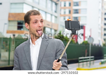 Man in formal clothing posing with selfie stick in urban environment showing tongue.