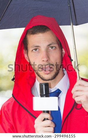 Successful handsome male journalist in red rain jacket working in rainy weather outdoors in park environment holding microphone and umbrella in live broadcasting.