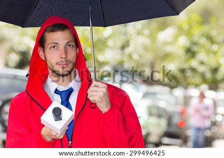 Successful handsome male journalist in red rain jacket working in rainy weather outdoors in park environment holding microphone and umbrella in live broadcasting.
