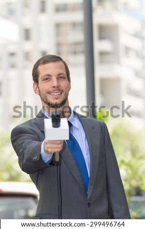 Attractive professional male news reporter wearing grey suit holding microphone, talking to camera from urban setting.