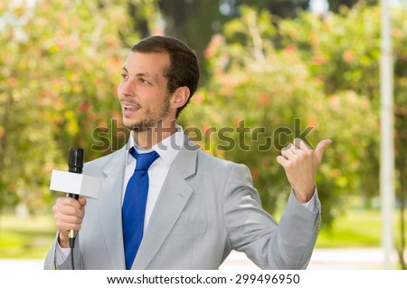 Successful handsome male news reporter in light grey suit working outdoors in park environment holding microphone in live broadcasting.