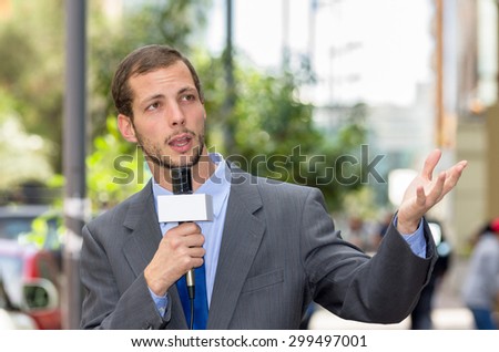 Attractive professional male news reporter wearing grey suit holding microphone, talking to camera from urban setting.