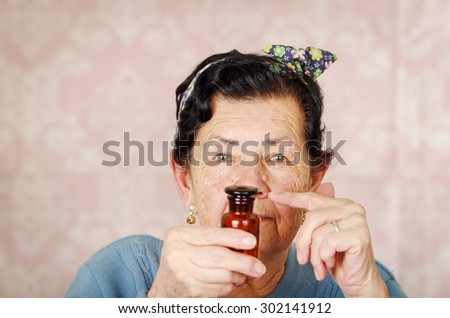 Older cool hispanic woman wearing blue sweater, flower pattern bow on head holding up a small red glass bottle for camera.