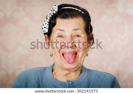Older cute hispanic woman wearing blue sweater and polka dot bowtie on head showing her tongue to the camera in front of pink wallpaper.