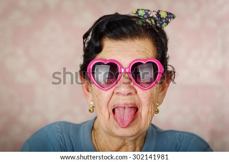 Older cool hispanic woman wearing blue sweater, flower pattern bow on head and pink heartshaped sunglasses looking into camera showing her tongue.