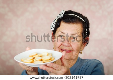 Older cute hispanic woman wearing blue sweater and polka dot bowtie on head holding up a plate of cookies.