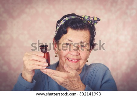 Older cool hispanic woman wearing blue sweater, flower pattern bow on head holding up a small red glass bottle and smiling to camera.