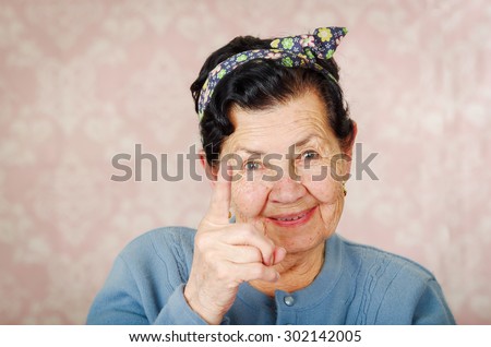 Older cute hispanic woman wearing blue sweater and flower pattern bow on head holding up one finger for the camera in front of pink wallpaper.