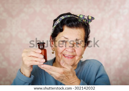 Older cool hispanic woman wearing blue sweater, flower pattern bow on head holding up a small red glass bottle and smiling to camera.