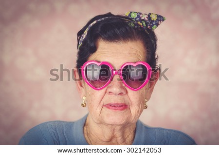 Older cool hispanic woman wearing blue sweater, flower pattern bow on head and pink heartshaped sunglasses looking into camera.