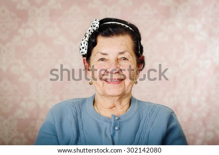 Older cute hispanic woman wearing blue sweater and polka dot bowtie on head smiling happily in front of pink wallpaper.