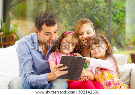 Family portrait of father, mother and two daughters sitting together in sofa playing happily with tablet.