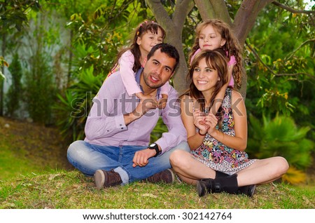 Family portrait of father, mother and two daughters sitting together in garden environment, children behind parents embracing them happily smiling.