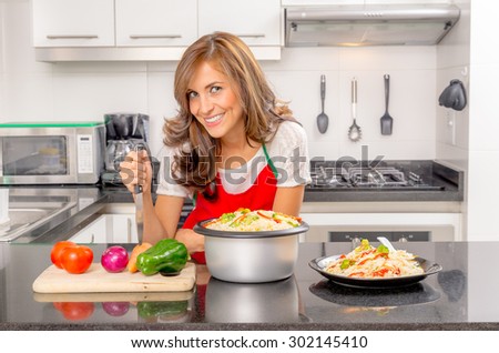 Hispanic beautiful woman cooking in modern kitchen by bench with veggies holding a knife down to carrot and smiling towards camera.