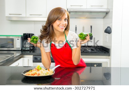 beautiful woman cooking in modern kitchen posing holding capsicum and smiling with plate of food on counter.